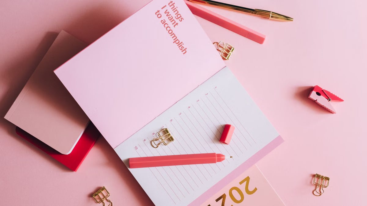 Organized workspace with pink planner, pen, and clips on pastel background.
